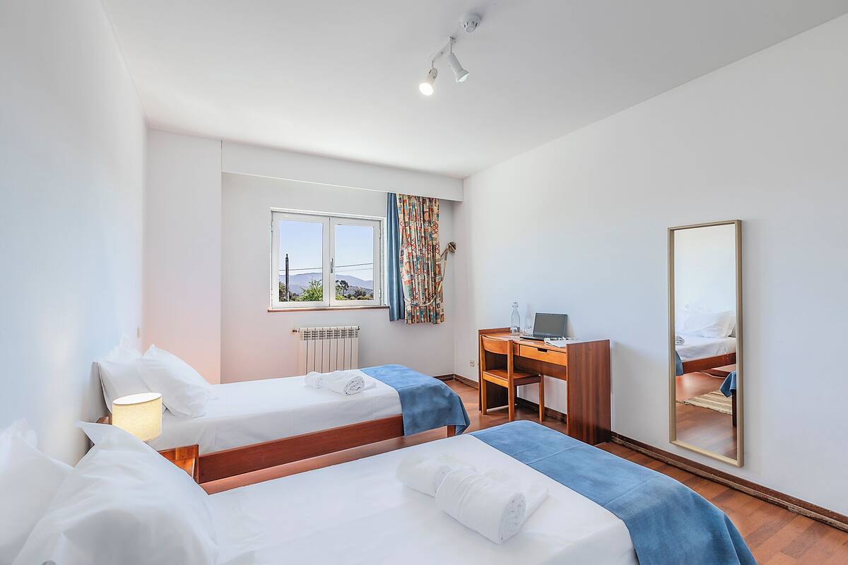A hotel room with two beds, a desk, a chair, a radiator, a window with curtains, and a mirror.