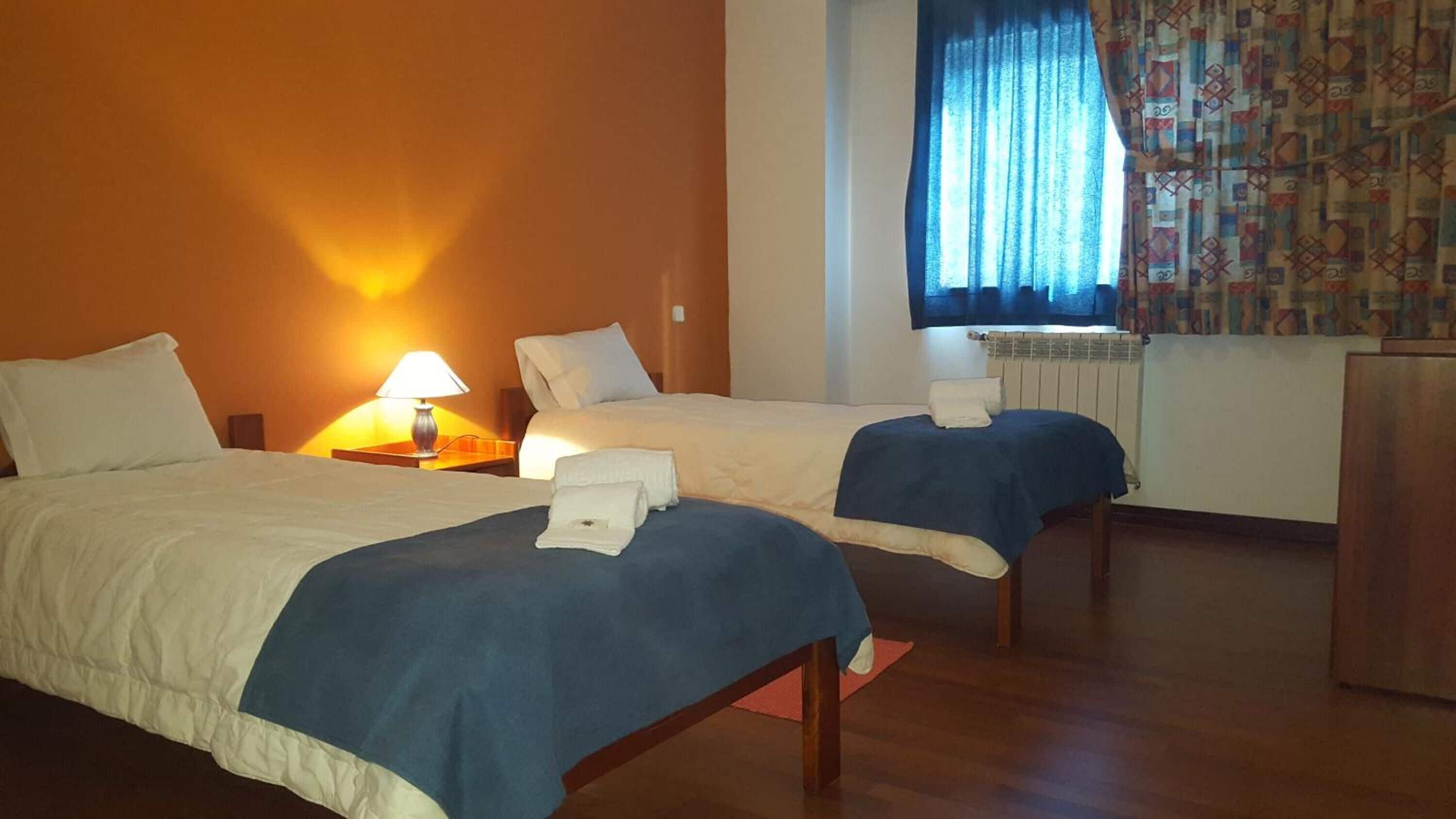 A hotel room with two beds, a nightstand with a lamp, a window with curtains, and a radiator.