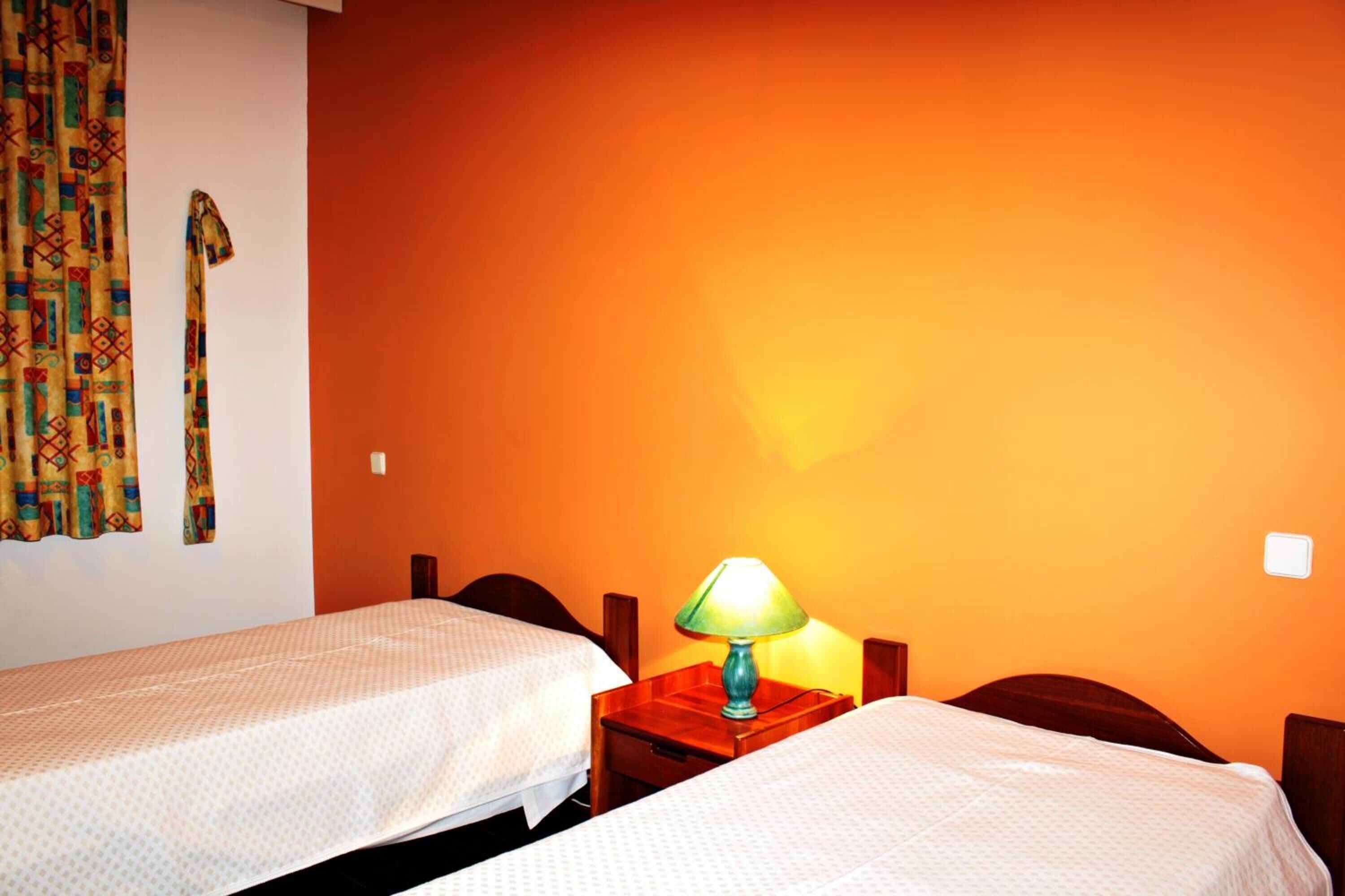 Une chambre d’hôtel avec deux lits simples, une table de chevet en bois avec une lampe et un rideau coloré.