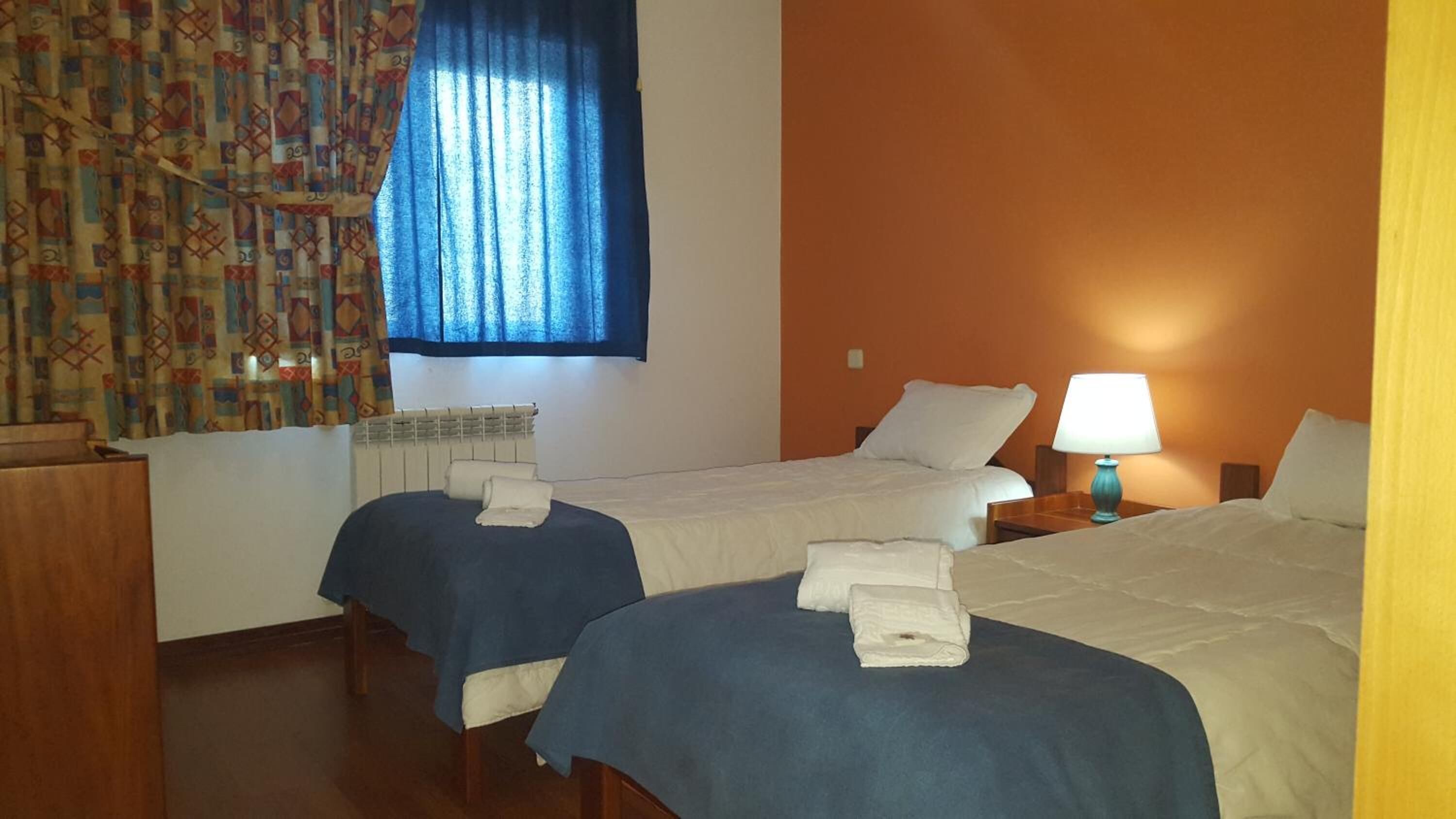 A hotel room with two beds, a window with blue curtains, a radiator, a bedside table with a lamp, and a wooden nightstand.