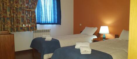 A hotel room with two beds, a window with blue curtains, a radiator, a bedside table with a lamp, and a wooden nightstand.