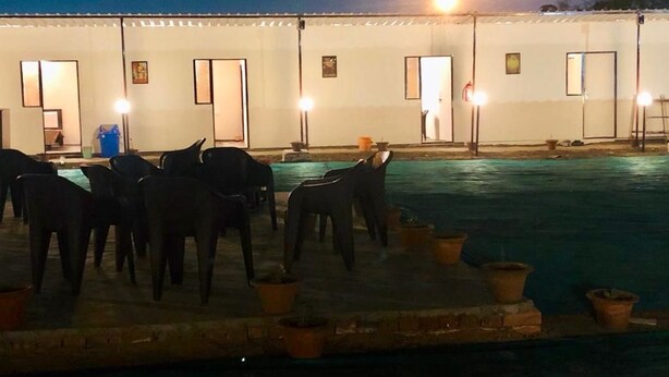 Un área junto a la piscina con sillas y plantas en macetas, un edificio con puertas y ventanas, y una farola.