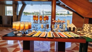 Mesa de buffet com diversos alimentos e bebidas, situada num espaço com vista para a água e uma escadaria.