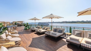 Um terraço com móveis de vime, almofadas listradas e guarda-sóis, com vista para um corpo d'água onde se vê um barco ao longe.