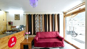 Zona de recepción de hotel con un mostrador de madera, un sofá rojo, cortinas estampadas y vistas al exterior a través de un gran ventanal.