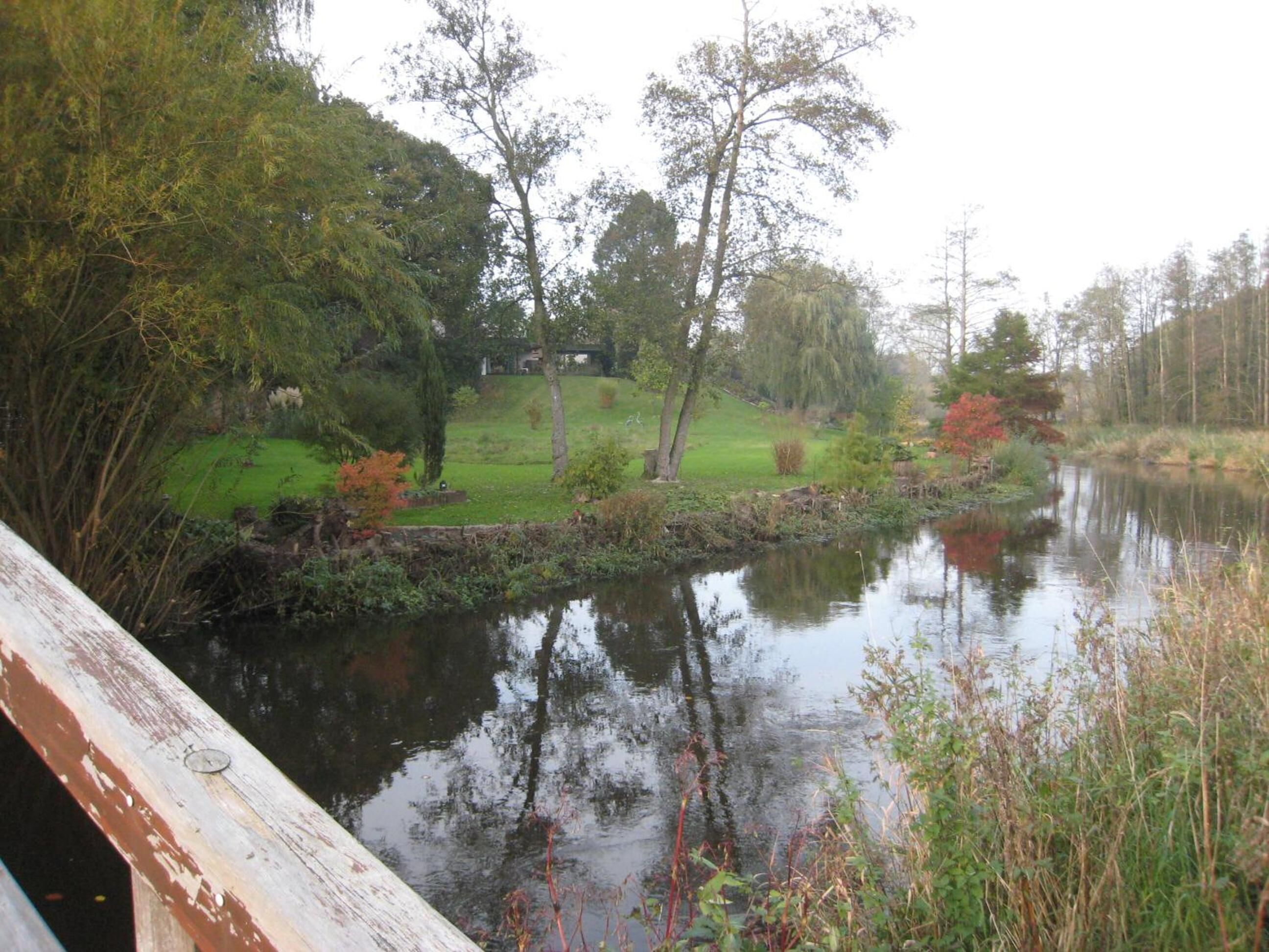 Eine friedliche Flusslandschaft mit einer Holzbrücke, üppiger Vegetation und einem Haus in der Ferne.
