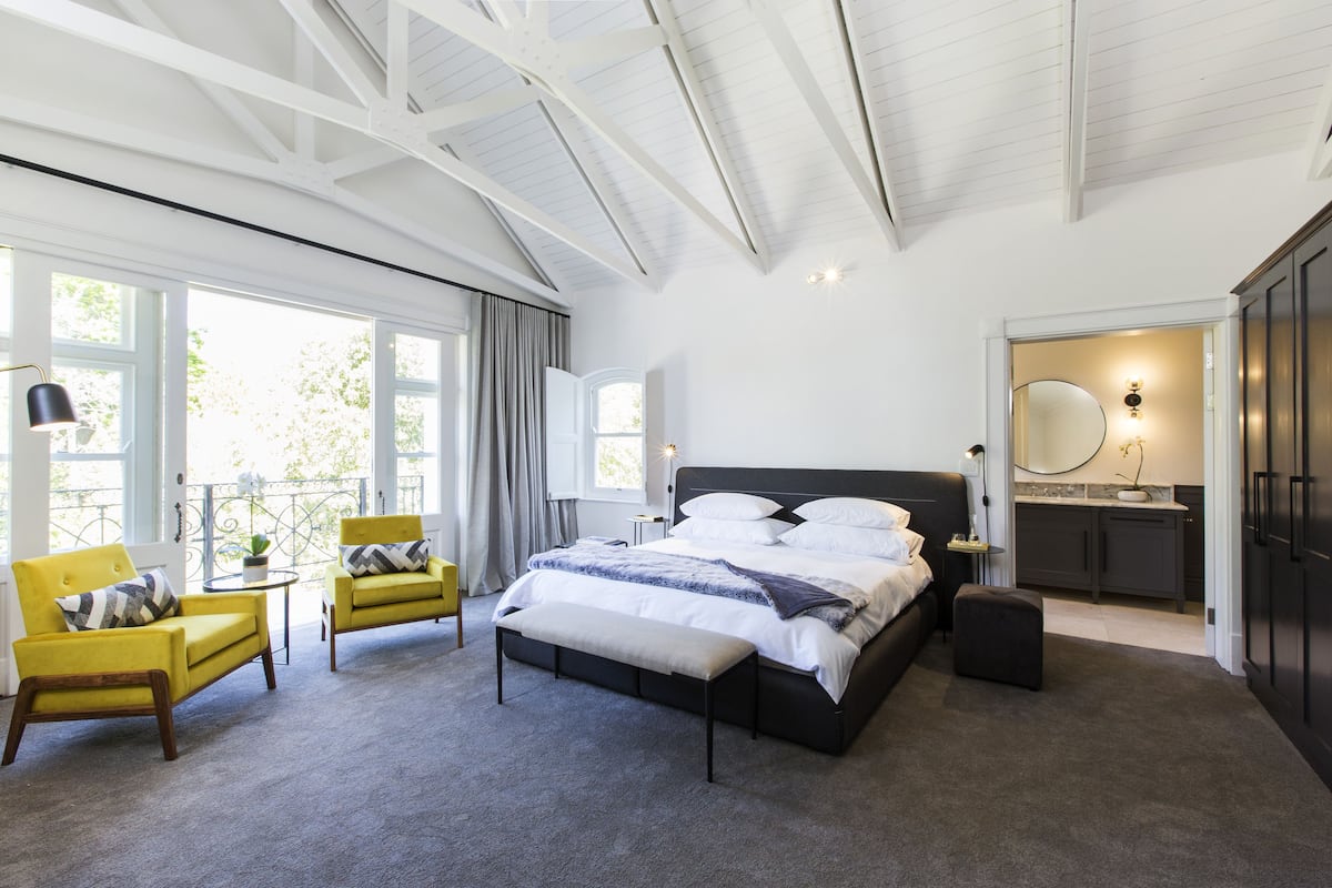 A bedroom with a large bed, two yellow armchairs, a bench, a small table with a lamp, and a view of the outdoors through large windows.