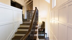 A grand staircase with a chandelier, white paneled walls, and a view to a room with a fireplace. - Arambrook Boutique Hotel (Cape Town)