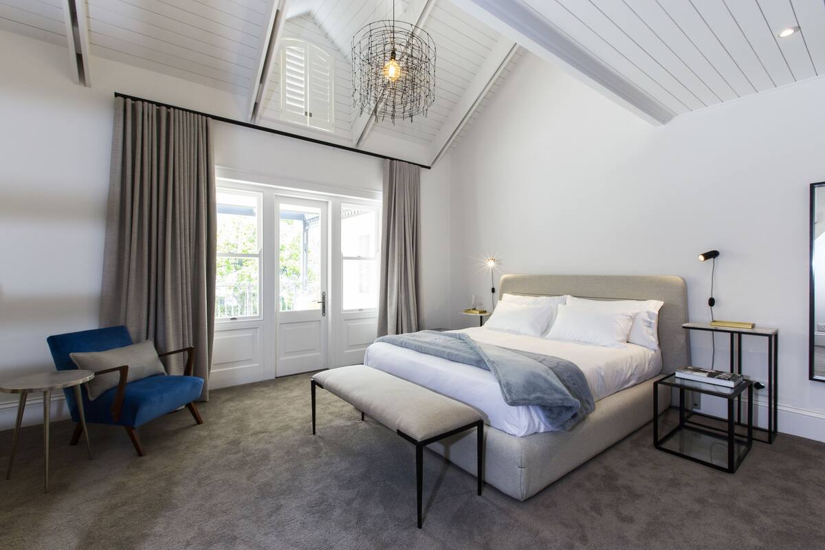 A bedroom with a large bed, a bench, a chair, a bedside table, and a hanging light fixture.