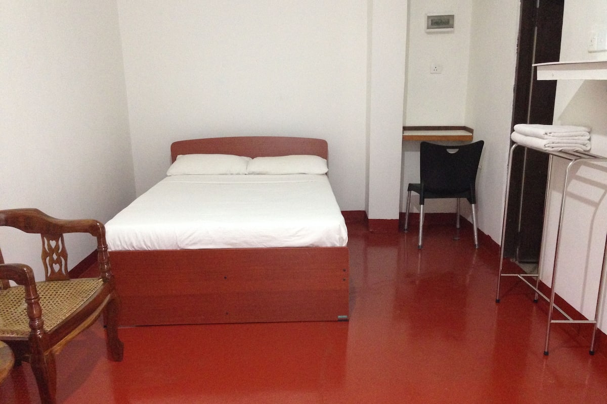A small, simple room with a bed, a wooden chair, a desk, and a radiator.