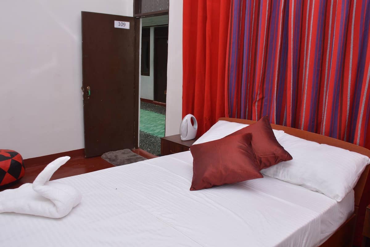 A hotel room with a bed, a pillow, a white towel, a red and black cushion, a white fan, and a door with a number sign.