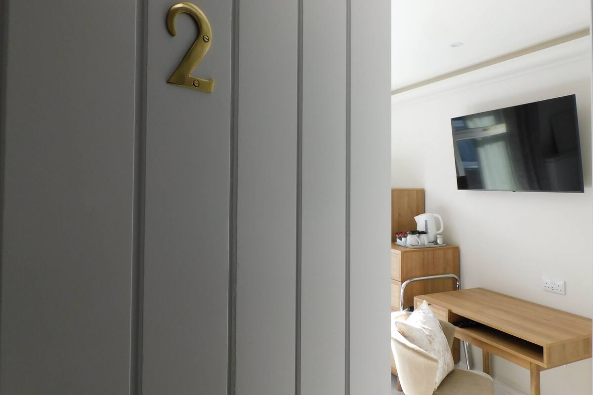 A modern hotel room with a grey door, a number '2' sign, a wooden desk, a chair, a TV mounted on the wall, and a small table with a kettle and cups.
