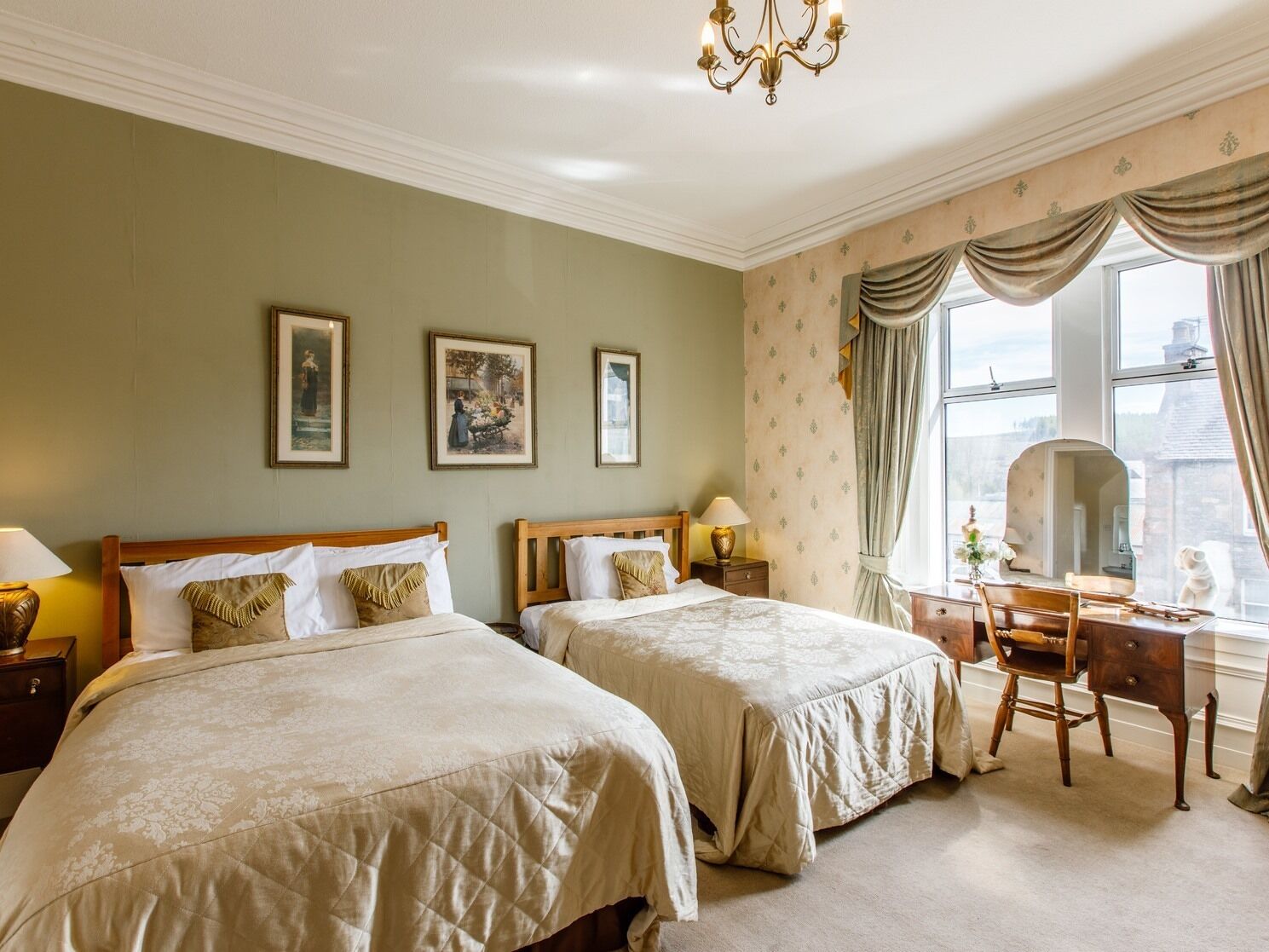 A hotel room with two beds, a desk, and a chair. There are framed pictures on the wall and a chandelier.