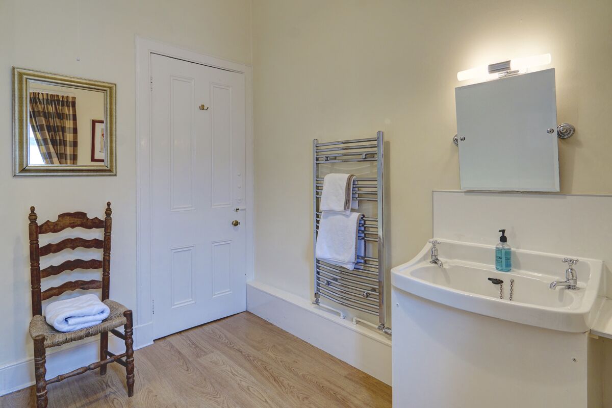A bathroom with a wooden chair, a white door, a towel rack, a sink with a mirror, and a framed picture on the wall.