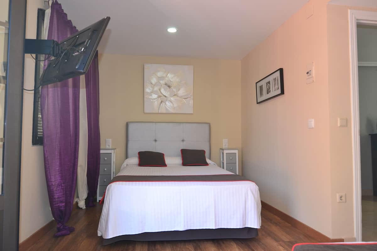 A bedroom with a bed, curtains, a nightstand, a wall-mounted artwork, and a door.