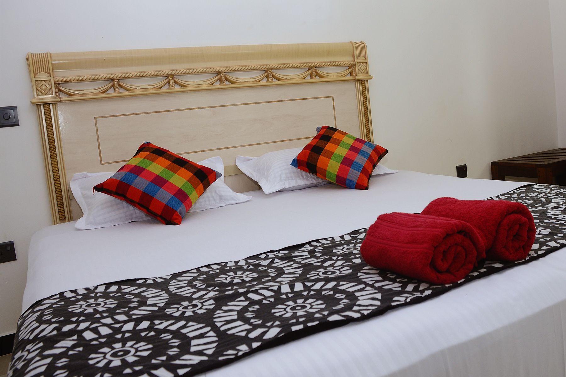 A neatly made bed with a patterned bedspread, two colorful pillows, and a rolled red towel.