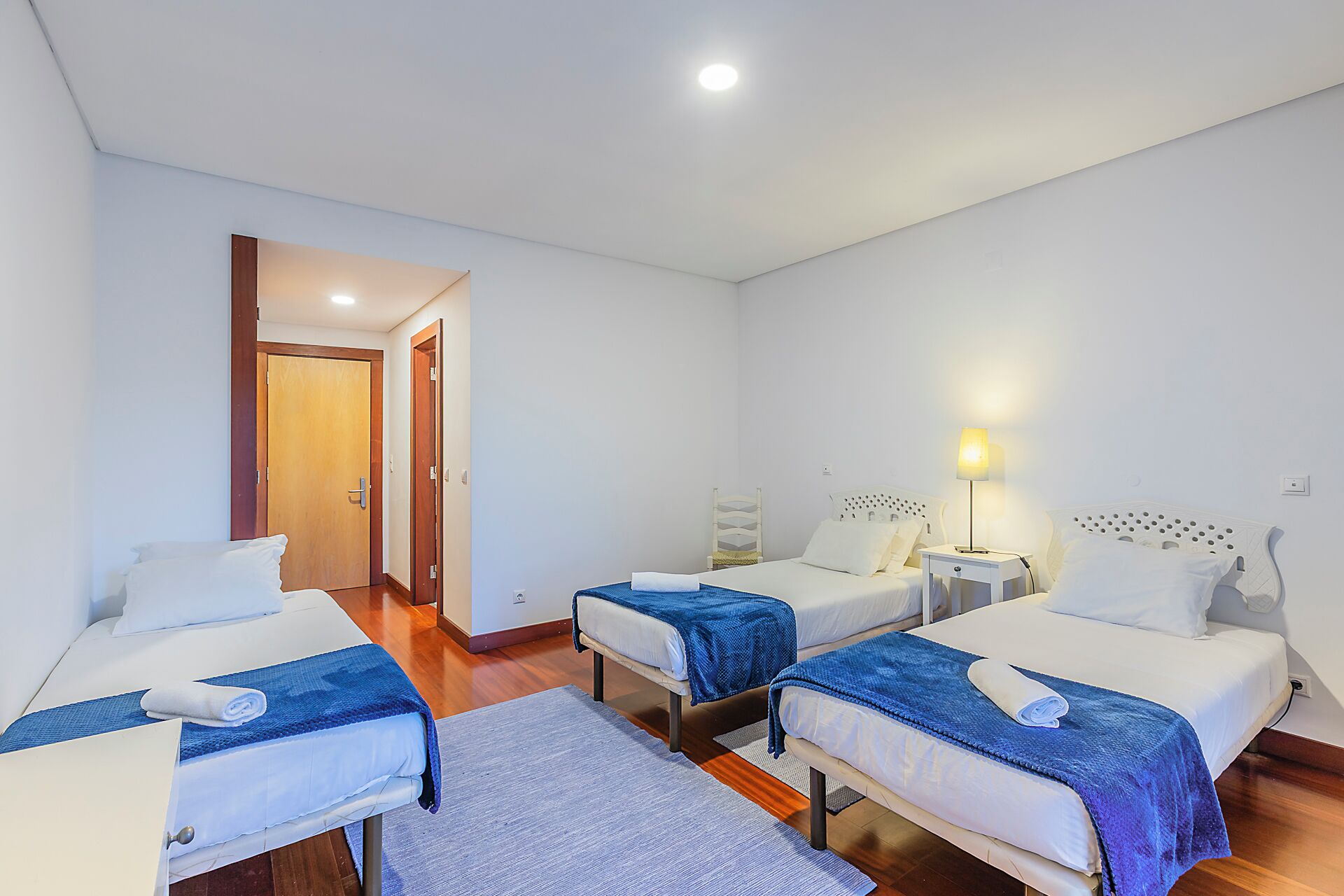 Quarto de hotel com três camas, piso de madeira e porta que dá para outro quarto.