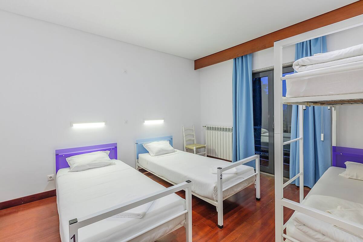 Une chambre d’hôtel avec deux lits simples, un lit superposé, une chaise, un radiateur et une fenêtre avec des rideaux.