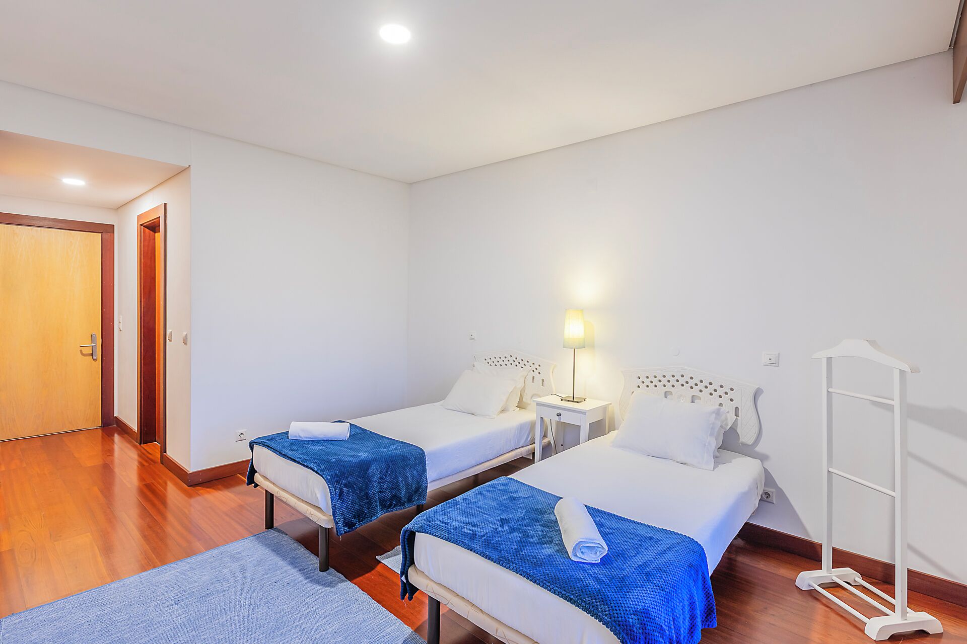 A hotel room with two beds, a nightstand with a lamp, a wooden floor, and a white wall.