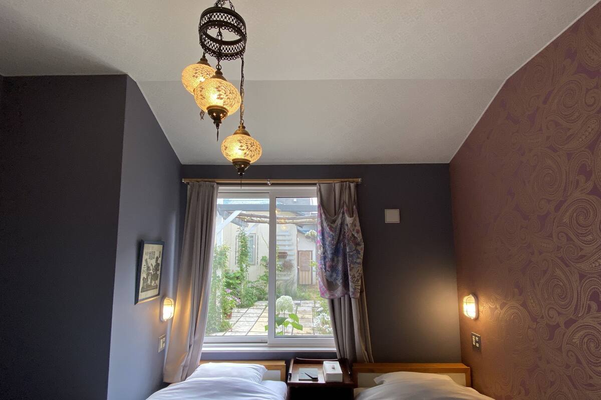 A bedroom with two beds, a window with curtains, a hanging light fixture, and a view of the outside garden.