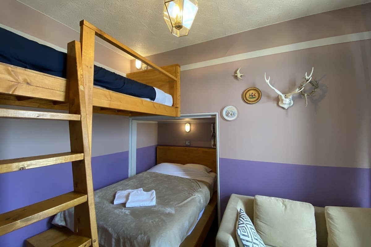 A bunk bed room with a wooden ladder, a bed with a white blanket, a chair with a patterned pillow, and wall decorations including a skull and antlers.