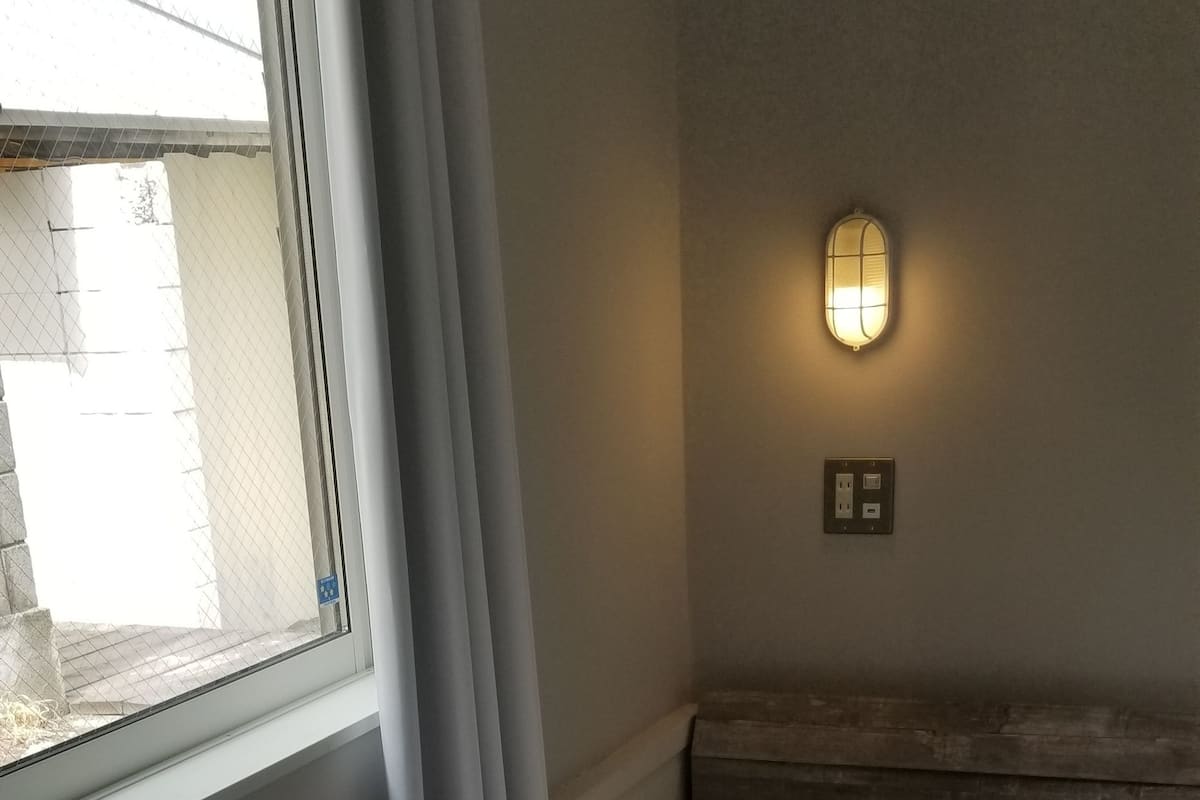 A neatly made bed with a quilted mattress, a pillow, and folded white towels. There is a window with curtains, a wall-mounted light fixture, and a small nightstand.