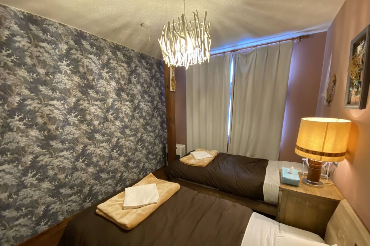 A hotel room with a large bed, a bedside table with a lamp, a window with curtains, and a wall with a floral wallpaper.