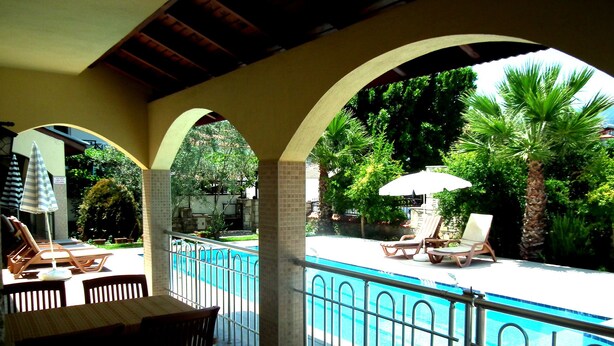 A pool area with lounge chairs and umbrellas, surrounded by a metal fence and greenery. - Villa Manzara (Ortaca)
