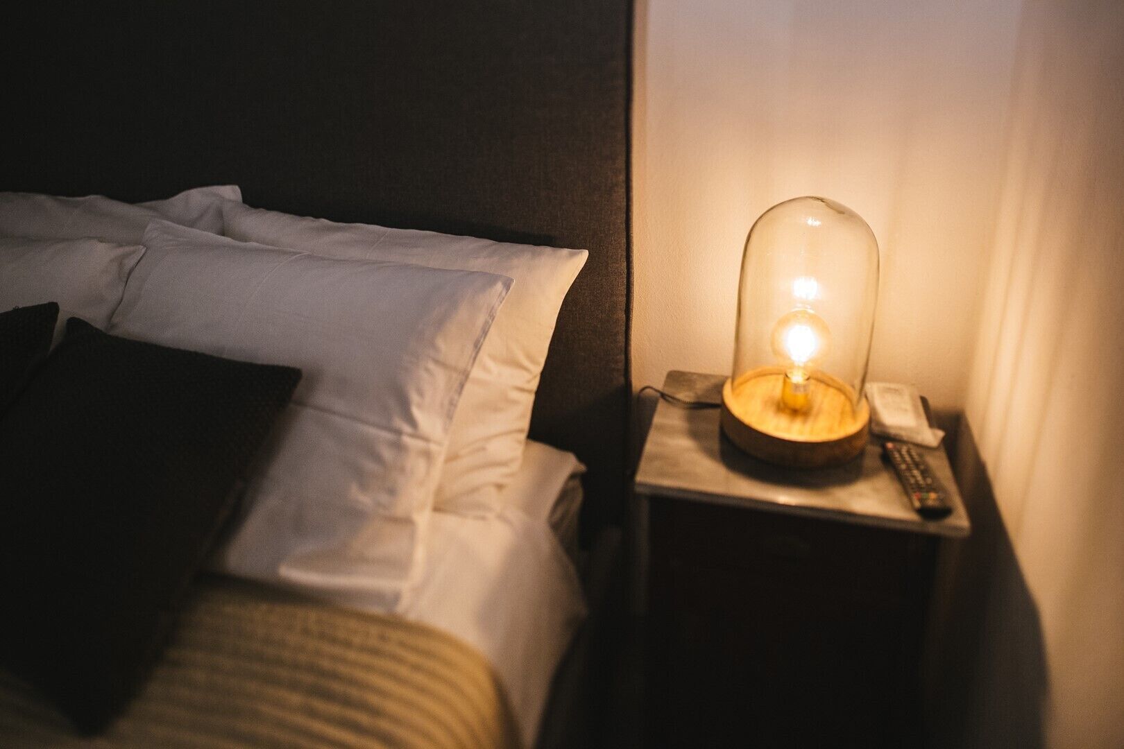 A bed with white linens and a black pillow, a bedside table with a lamp, and a remote control.