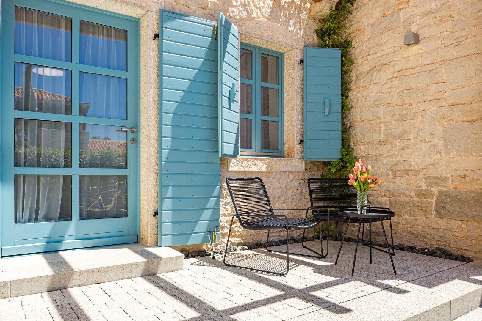 Un edificio in pietra con persiane blu, un piccolo patio con due sedie e un tavolo, e una finestra con una bicicletta all'interno.