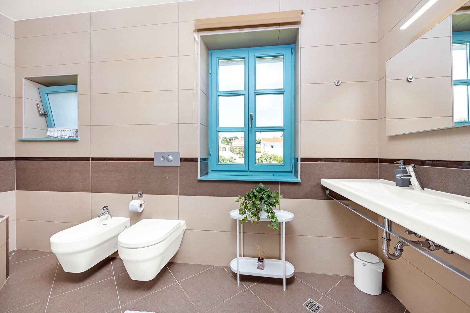Et moderne badeværelse med toilet, bidet, håndvask og et vindue med blå skodder.