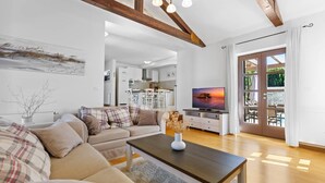 A modern living room with a sofa, coffee table, and TV. There's a kitchen in the background and a view of the outdoors through a glass door.