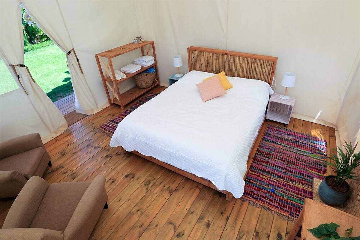 A bedroom with a wooden bed, two bedside tables, a shelf with towels, a potted plant, and a view of a green outdoor area.