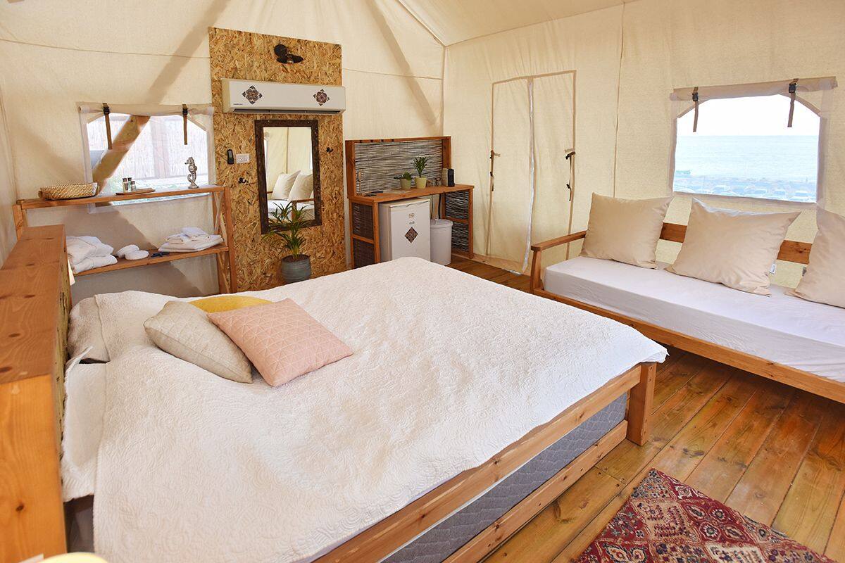 A tented accommodation with a large bed, a smaller bed, a wooden desk, a mirror, and a view of the sea.