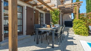 Un patio exterior con mesa lista para comer, pérgola de madera, paredes de piedra y una zona de piscina.