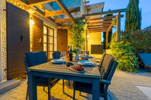 An outdoor dining area with a table set for two, surrounded by chairs, and a pergola overhead.
