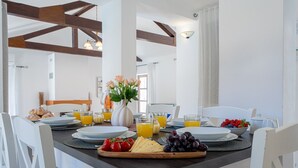 Una escena de desayuno con una mesa preparada para cuatro personas, incluyendo platos, vasos de jugo de naranja, un florero y una tabla con queso, tomates y uvas.
