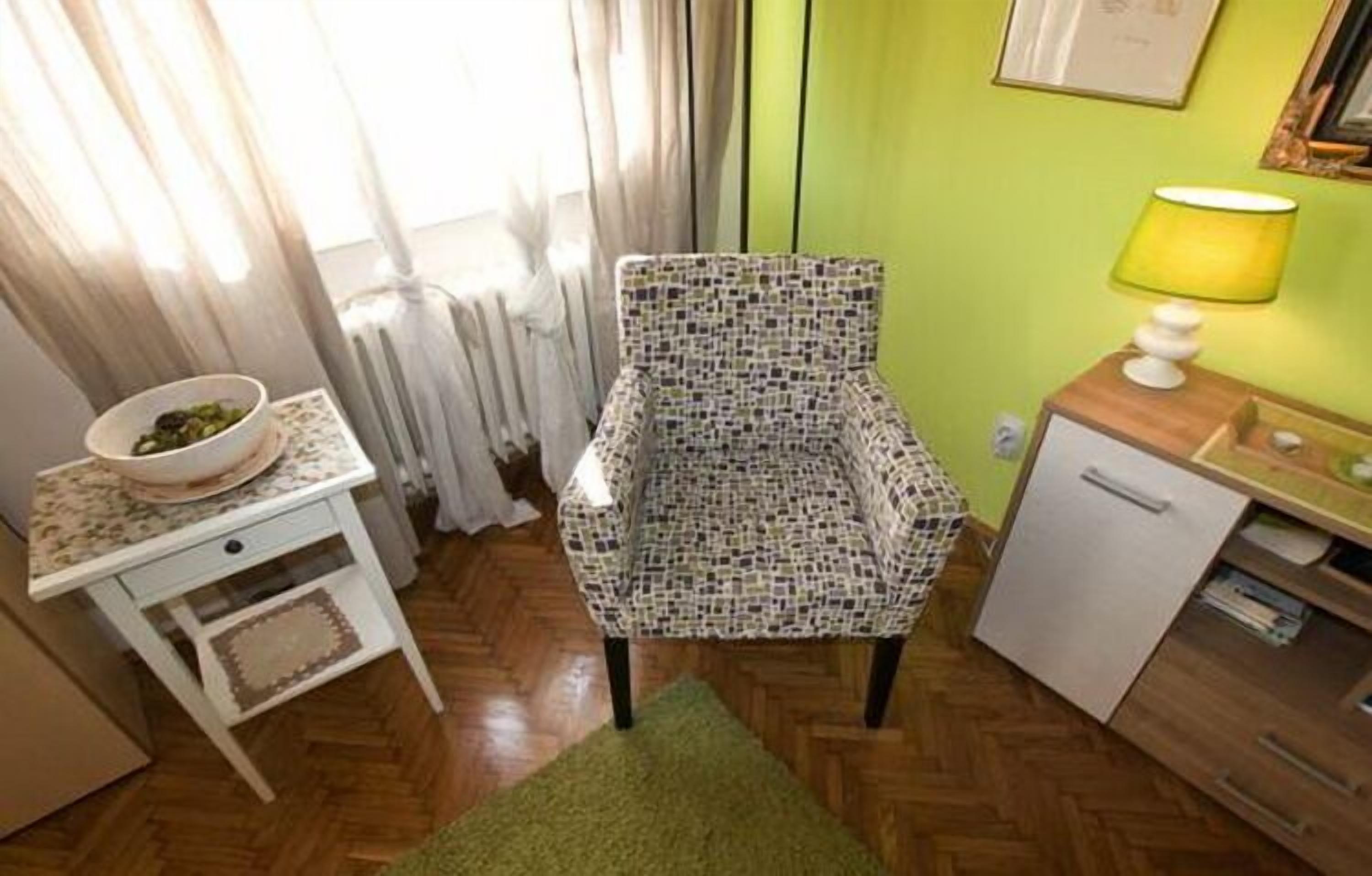 A small, compact room with a patterned chair, a small table with a bowl, a lamp, and a compact refrigerator.