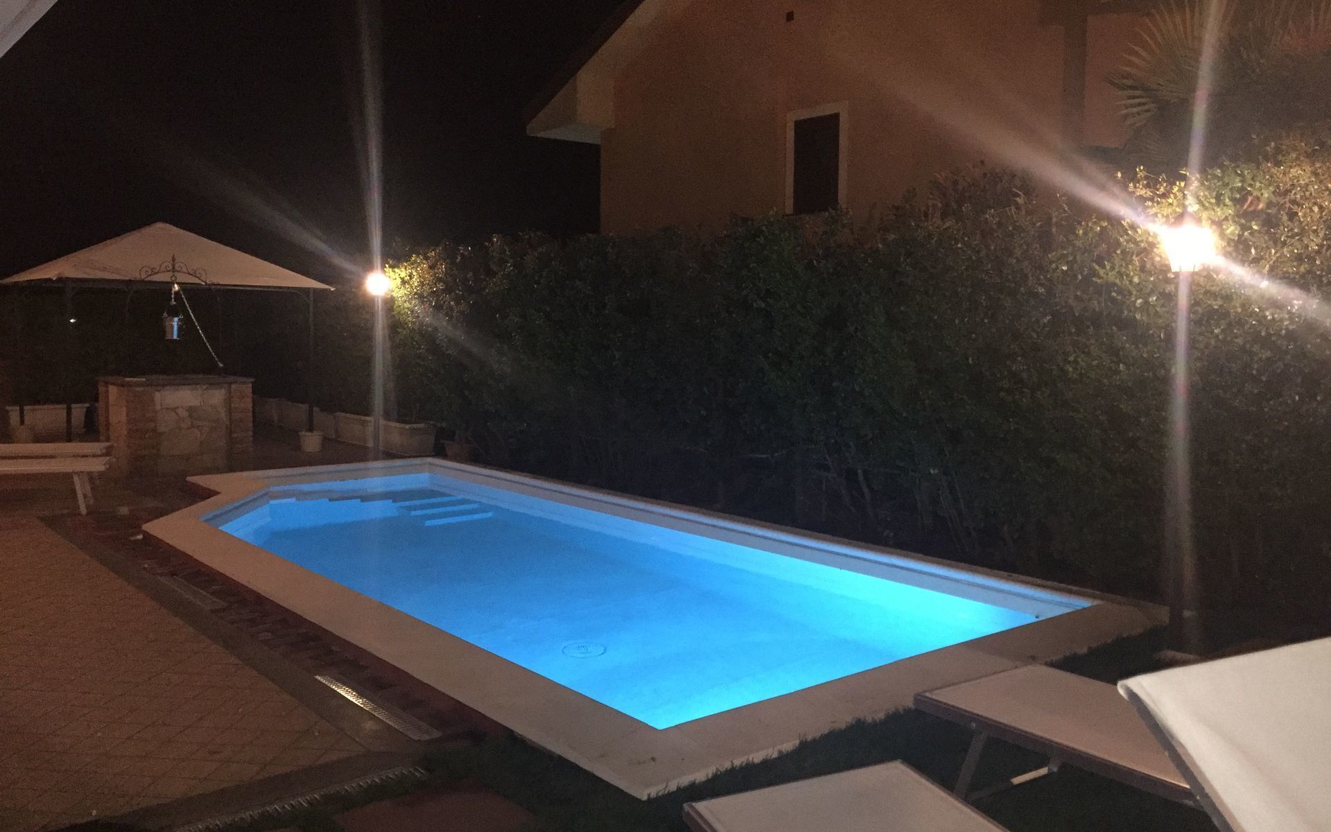 A rectangular swimming pool with a blue tint, surrounded by a stone wall and a hedge. There are two outdoor lights on either side of the pool.