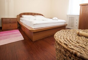 Uma cama bem arrumada com lençóis brancos, uma mesa de cabeceira de madeira com gavetas, um banquinho de vime e um tapete listrado rosa.