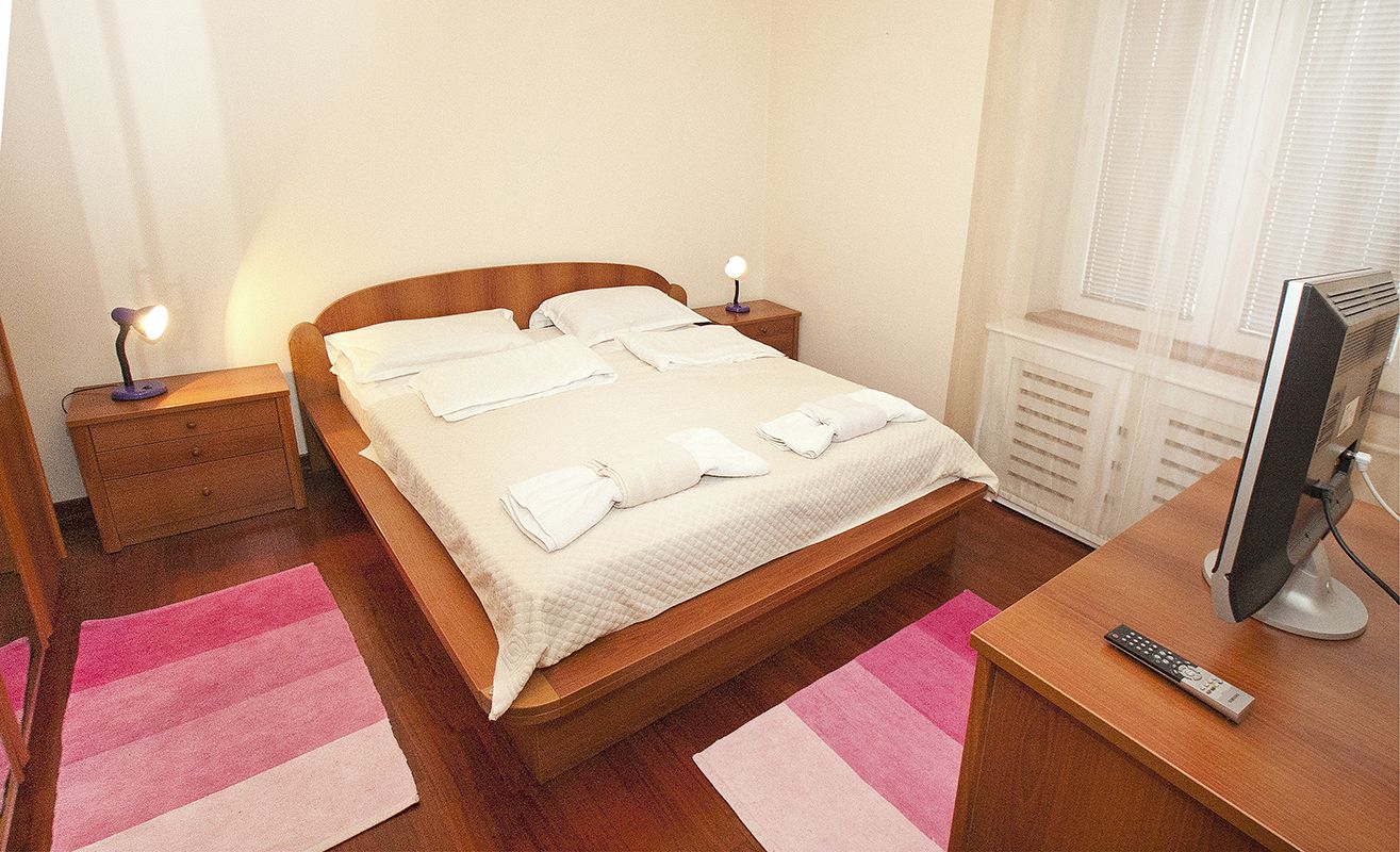 A neatly made bed with white linens and towels, a bedside lamp, a wooden nightstand, a television on a wooden table, and a pink and white striped rug.