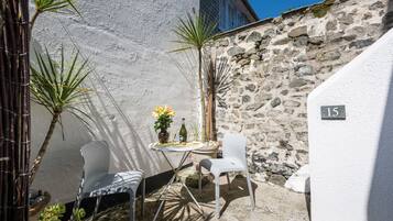 Un pequeño espacio al aire libre con dos sillas blancas, una mesa con un florero y una botella de vino. Se ve una pared de piedra y una pared blanca con el número '15'.