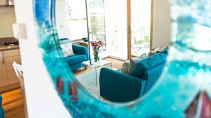 Uma sala de estar moderna com um sofá azul, uma mesa de centro de vidro e vista para o exterior através de uma porta de vidro.