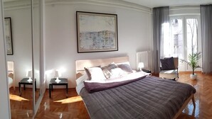 A bedroom with a large bed, a chair, a small table, and a potted plant. There is a window with curtains and a view of the outside. - Apartment Art Galery (Belgrade)