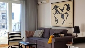 A modern living room with a grey sofa, striped armchair, and a coffee table. A large abstract painting hangs on the wall, and there is a window with curtains. - Apartment Art Galery (Belgrade)