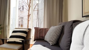 A modern living room with a sofa and armchair, a window with curtains, and a view of buildings outside. - Apartment Art Galery (Belgrade)