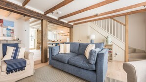 Un salon avec un canapé bleu, un fauteuil beige et un tapis à motifs. On y trouve un escalier avec une rampe en bois et un mur blanc orné d’un élément décoratif.