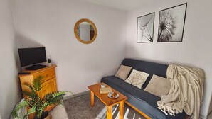 A living room with a sofa, a coffee table, a TV on a wooden cabinet, a mirror, and two wall art pieces. - Charming Studio for 4 with Balcony, Parking & Ski Storage – Near Ski Resorts (Luz-Saint-Sauveur)