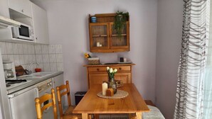 A compact kitchen with a microwave, stove, and sink. There is a wooden dining table with chairs and a bench. A cabinet with glass doors and a plant is above the table. - Charming Studio for 4 with Balcony, Parking & Ski Storage – Near Ski Resorts (Luz-Saint-Sauveur)