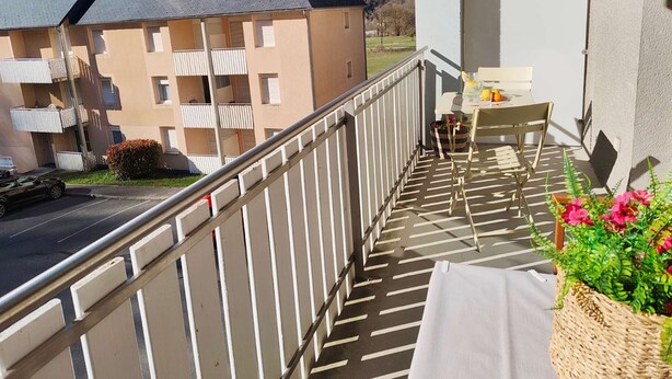 A balcony with a table and chairs, a wicker basket with flowers, and a view of a building and mountains. - Charming Studio for 4 with Balcony, Parking & Ski Storage – Near Ski Resorts (Luz-Saint-Sauveur)
